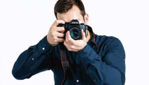 A professional photographer demonstrating the proper two-handed grip technique with a right hand on the camera and left hand supporting the lens for stability.