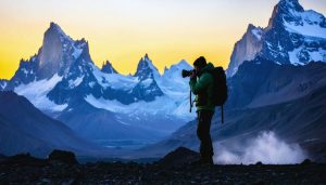 Silhouette of an exploration photographer with weather-resistant camera equipment capturing a sunrise over snowy mountain peaks.