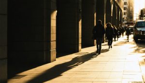 Street photograph at golden hour showcasing intersecting natural and artificial lines with commuters moving amidst long shadows and architectural elements as frames.