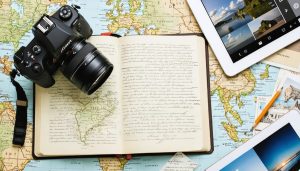 A camera placed on an open travel journal with handwritten notes and sketches, next to a digital tablet displaying a photo journal app, representing the blend of analog and digital journaling for travel photographers.